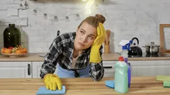 Unavená žena s úklidovými prostředky v kuchyni, opřená o stůl s rukavicemi.